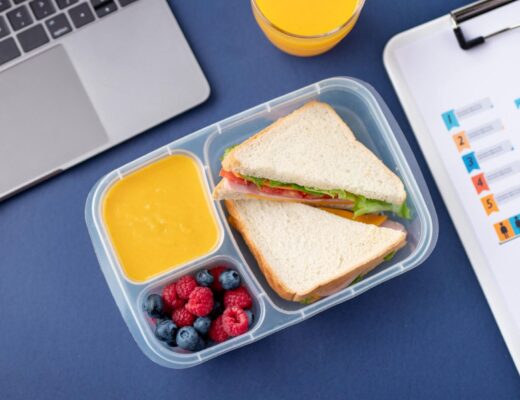 Lunch w plastikowym pojemniku podzielonym na trzy przegródki: kanapki z szynką, serem, sałatą i pomidorem, porcja malin i borówek oraz kremowa zupa lub sos w kolorze żółtym