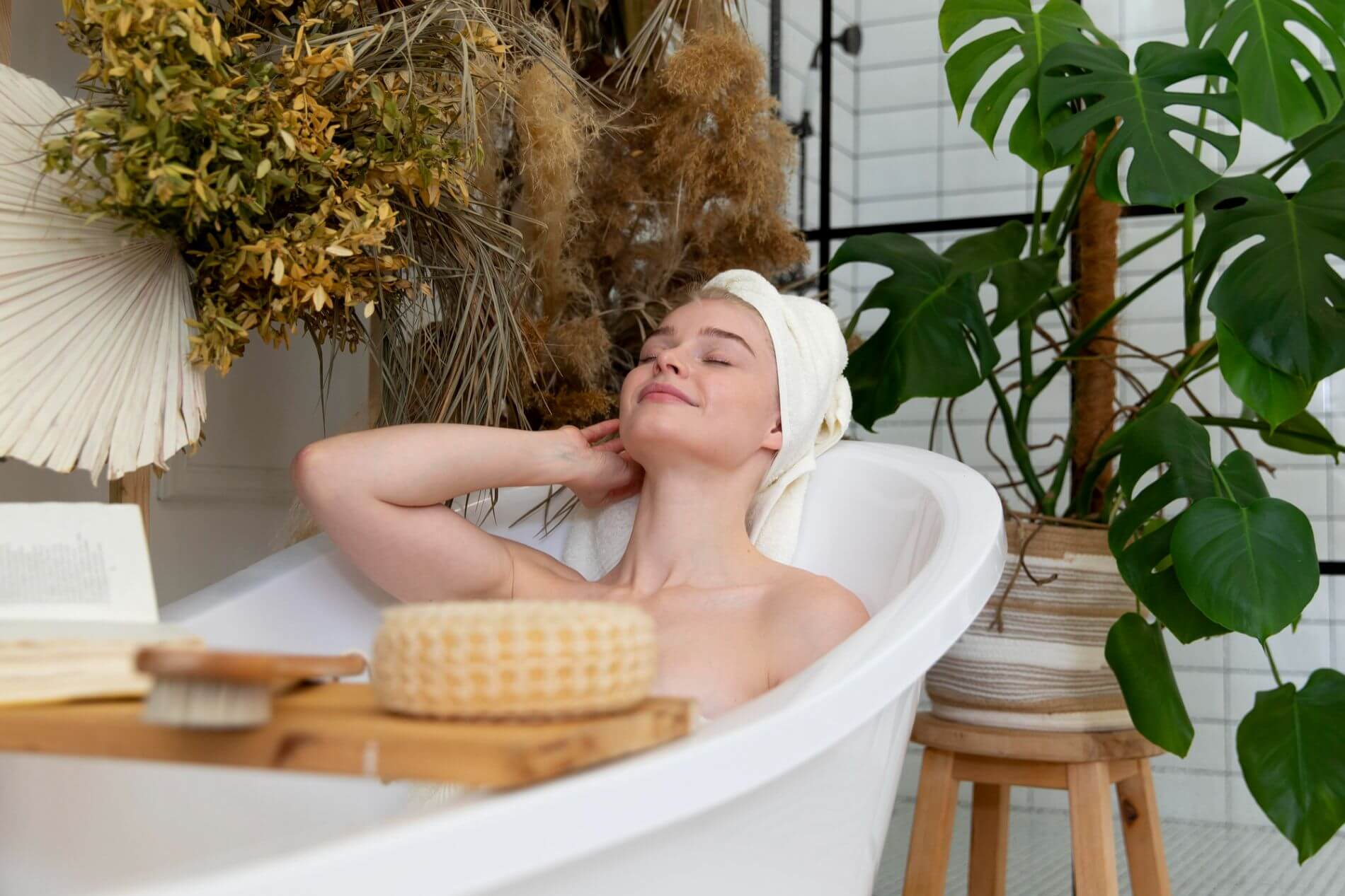 Kobieta odpoczywa w wannie z ręcznikiem na głowie, otoczona roślinami i akcesoriami do pielęgnacji, czyli jak zrobić domowe spa
